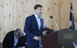 A man in a blue suit speaks into a microphone at a podium labeled "Dillon County Chamber of Commerce," with an American flag visible in the background.