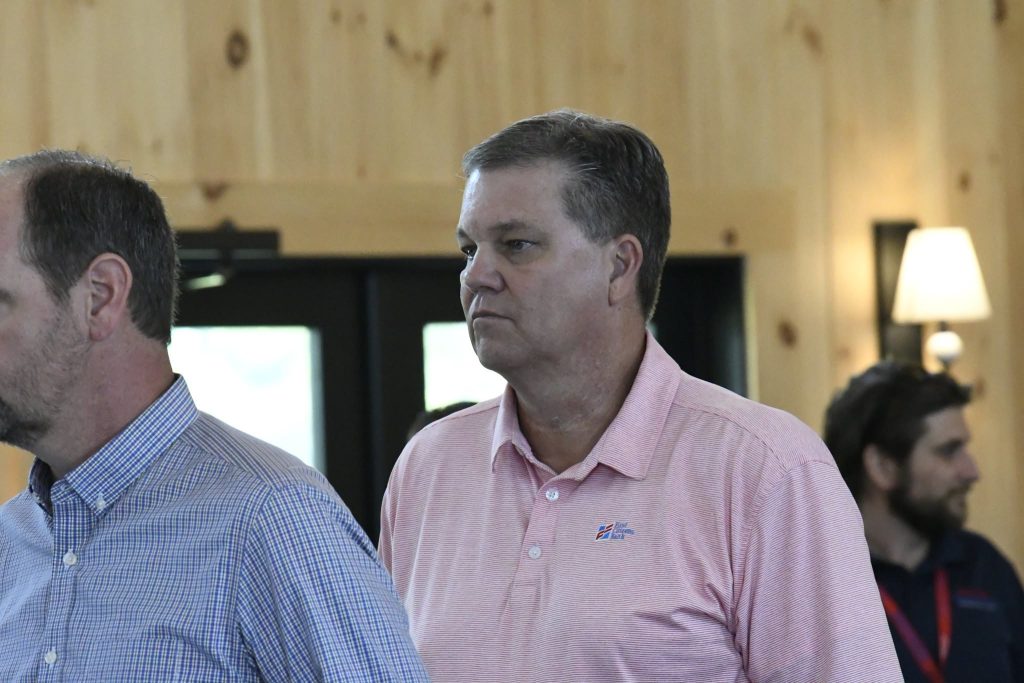 Two men standing indoors, one in a pink shirt and the other in a blue checkered shirt, with people and wooden walls in the background.