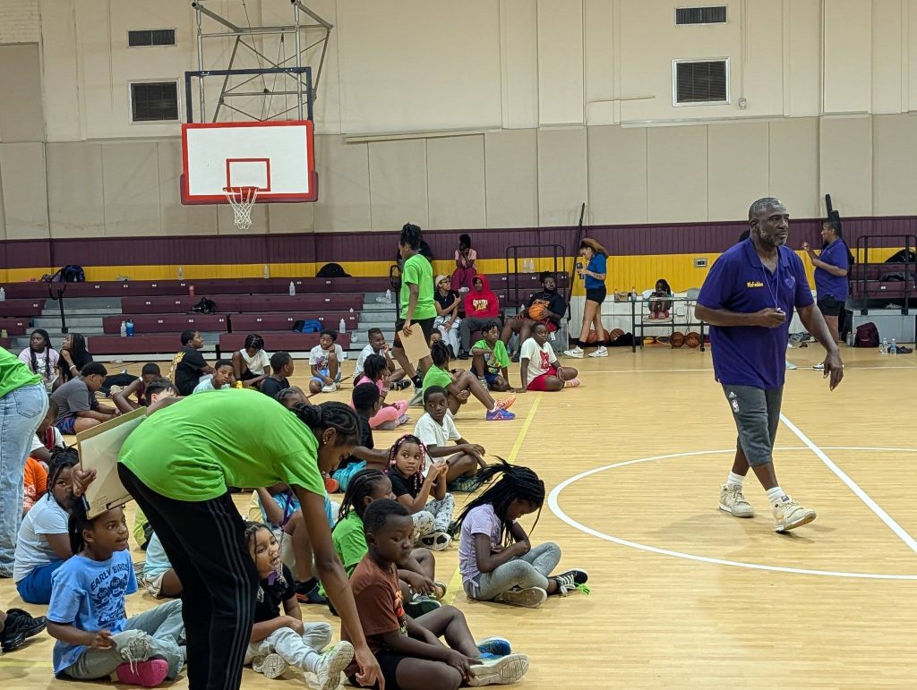 Children sitting on a gym floor with adults leading activities; a basketball hoop and bleachers are visible in the background.