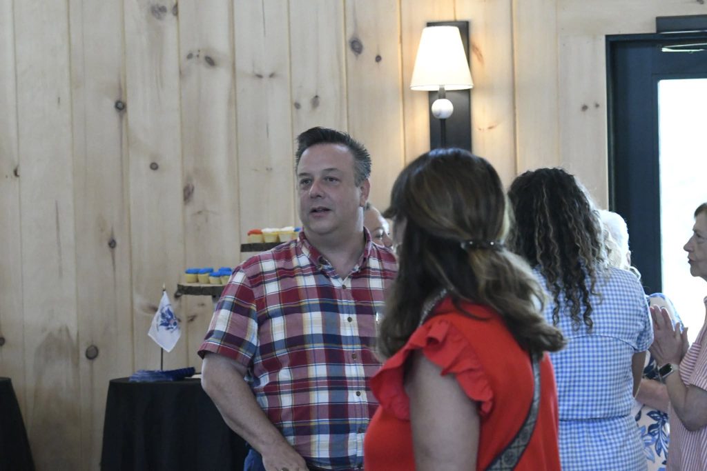 A man in a plaid shirt stands indoors at a gathering, with people talking in the background and cupcakes displayed on a table.