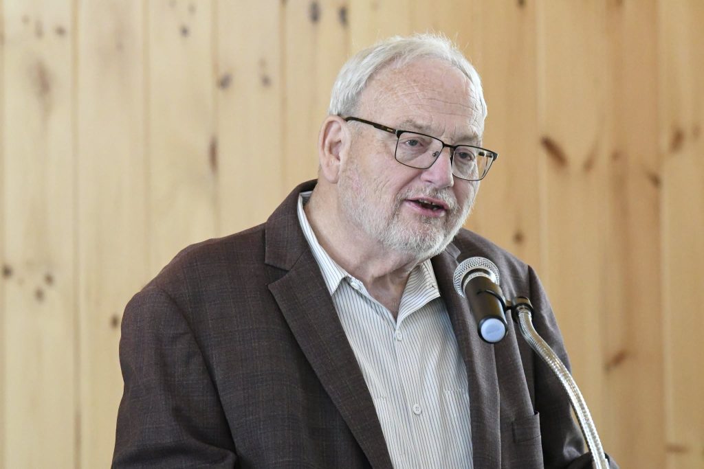 older man with glasses and a beard speaks into a microphone, wearing a brown jacket and striped shirt, in front of a wooden wall.