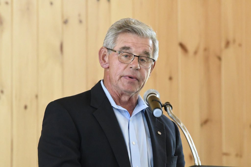 Older man with gray hair and glasses speaking at a microphone, wearing a suit jacket and dress shirt, with a wooden wall background.
