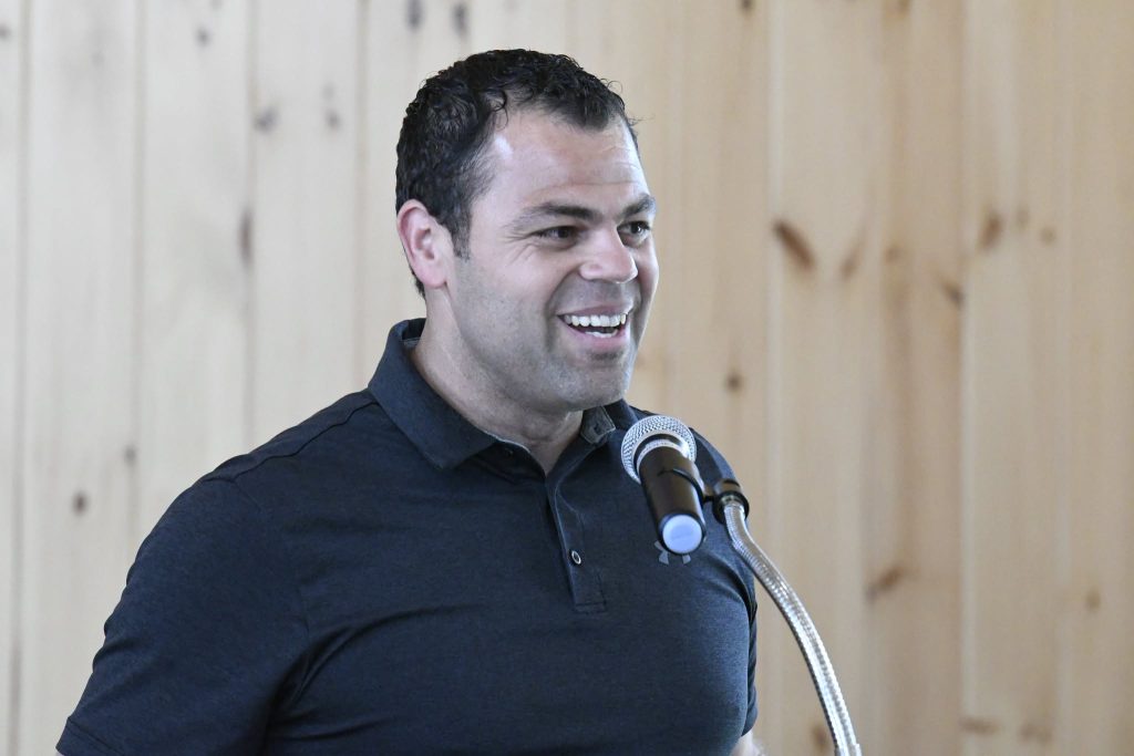 A man in a dark polo shirt smiles while speaking at a microphone, standing in front of a light wooden wall.