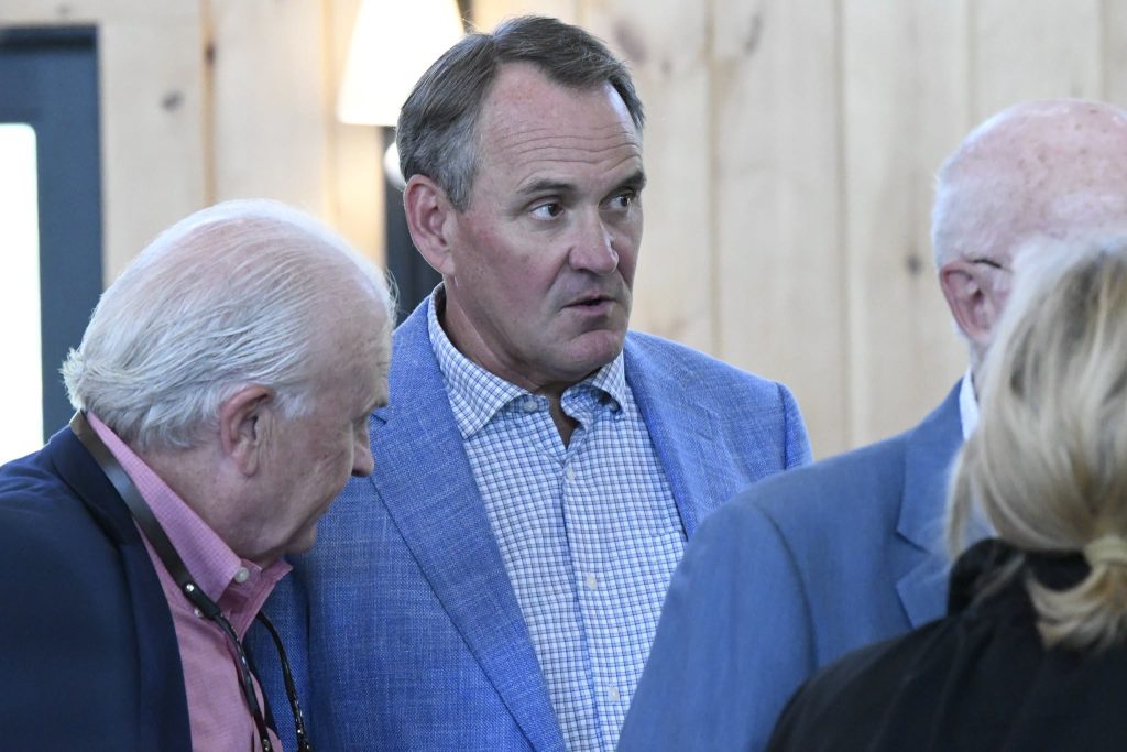 A man in a blue blazer talks with a group of people indoors, appearing engaged in conversation.