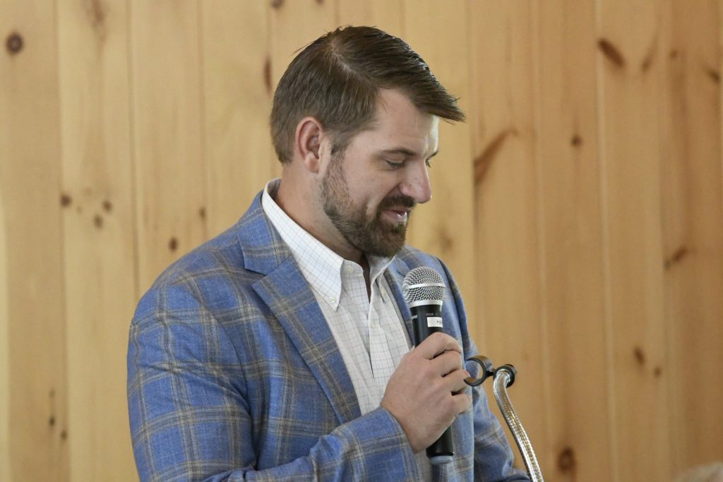 Man in a blue plaid jacket speaking into a microphone, standing in front of a wooden wall.