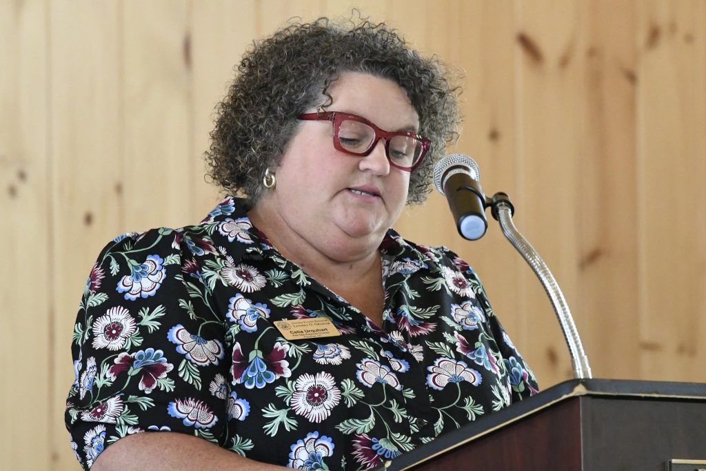 Woman with curly hair and glasses speaks at a podium with a microphone, wearing a floral blouse and a name tag, against a wooden background.