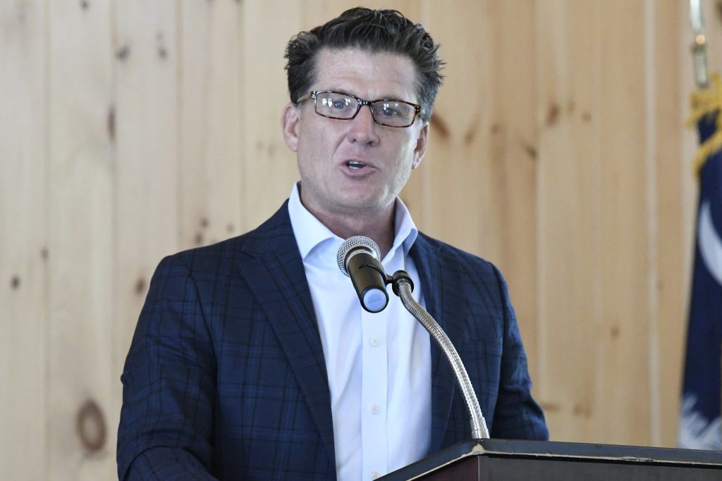 A man wearing glasses and a suit jacket speaks at a podium with a microphone, with a wooden wall and partial flag in the background.