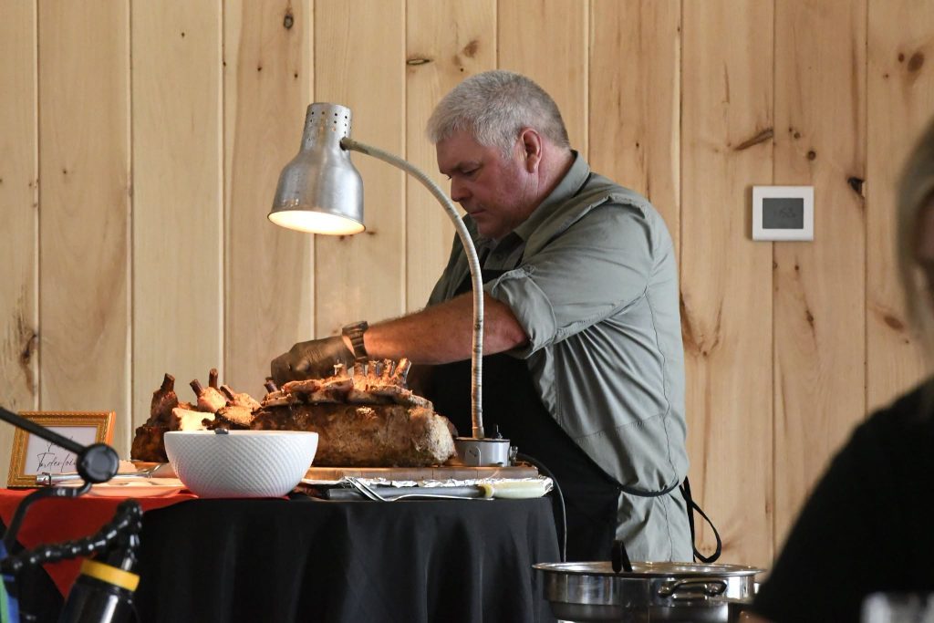 A man carving a large rack of roasted meat under a heat lamp at a buffet table, with a bowl and utensils nearby.