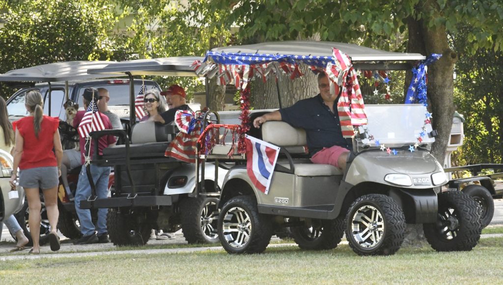 People gather near golf carts decorated with American flags and red, white, and blue streamers, celebrating outdoors under trees on a sunny day.