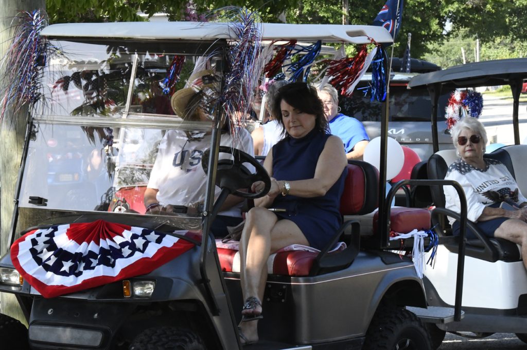 People ride decorated golf carts with patriotic red, white, and blue banners and tinsel, likely during an Independence Day parade or celebration.