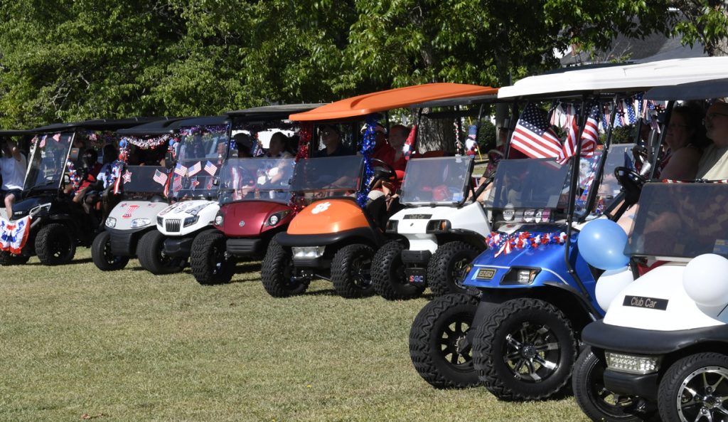 A row of decorated golf carts with American flags and festive banners parked on grass, people inside celebrating outdoors on a sunny day.