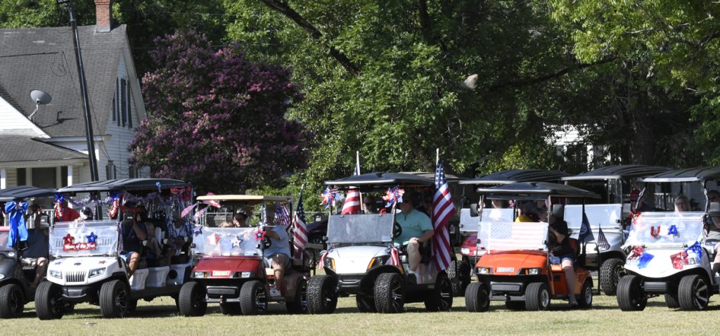 Golf carts decorated with American flags and patriotic colors line up on grass, with people sitting inside, celebrating an outdoor event in a residential neighborhood.