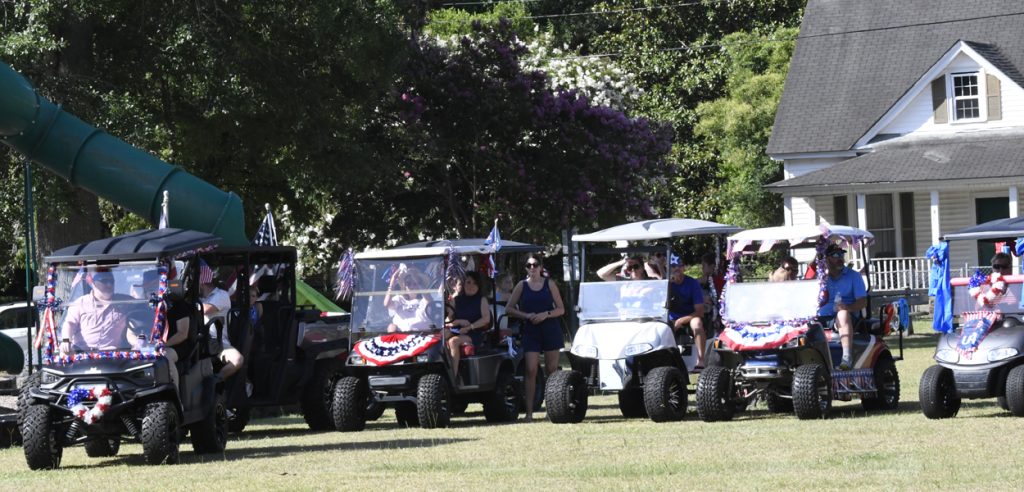 People in golf carts decorated with red, white, and blue for a patriotic event, gathered near a house and park area on a sunny day.
