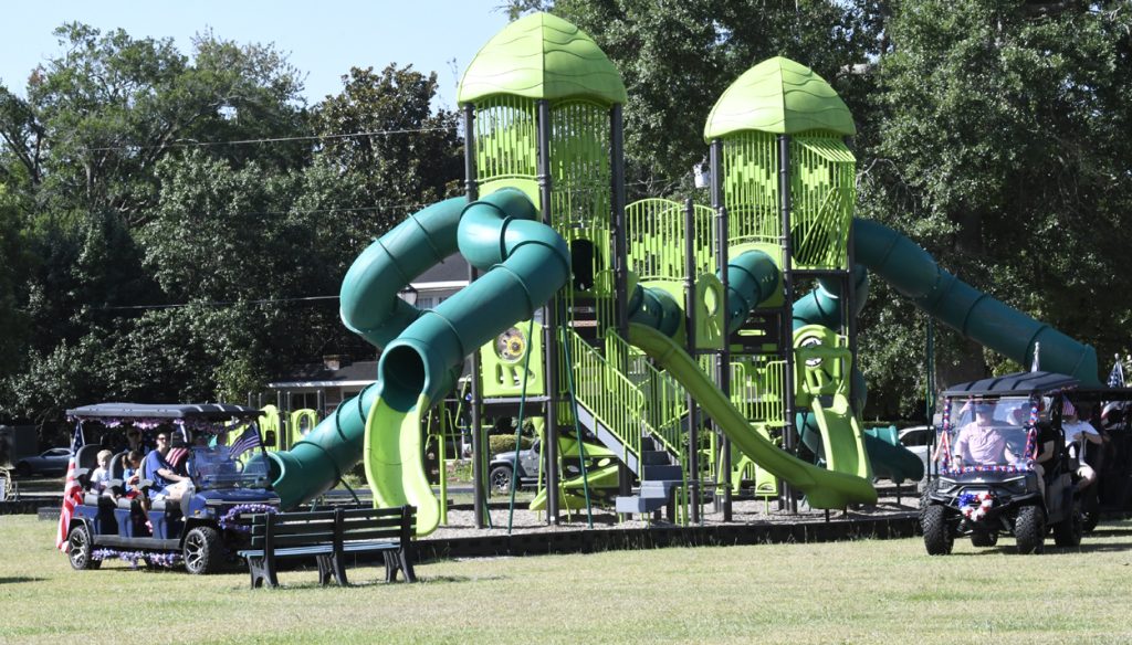 Large green playground structure with twisty slides in a park, two golf carts with people parked nearby, surrounded by trees and grass on a sunny day.