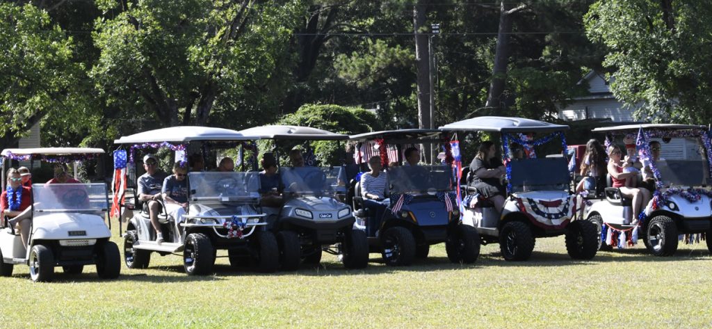 Golf carts decorated with American flags and patriotic bunting, people sitting inside, parked on grass, likely at a festive community event or parade.