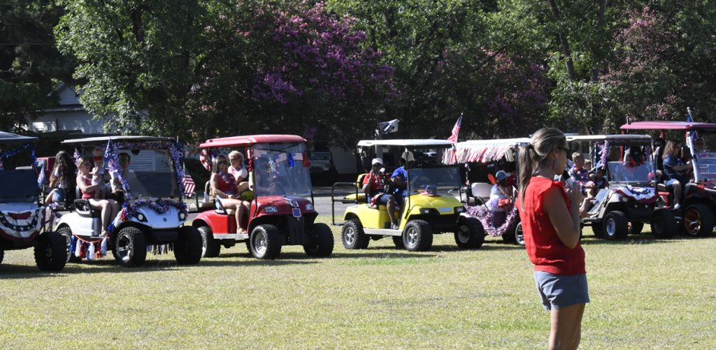 People sitting in decorated golf carts during a sunny outdoor event, with trees and purple flowers in the background. A woman in red stands in the foreground.