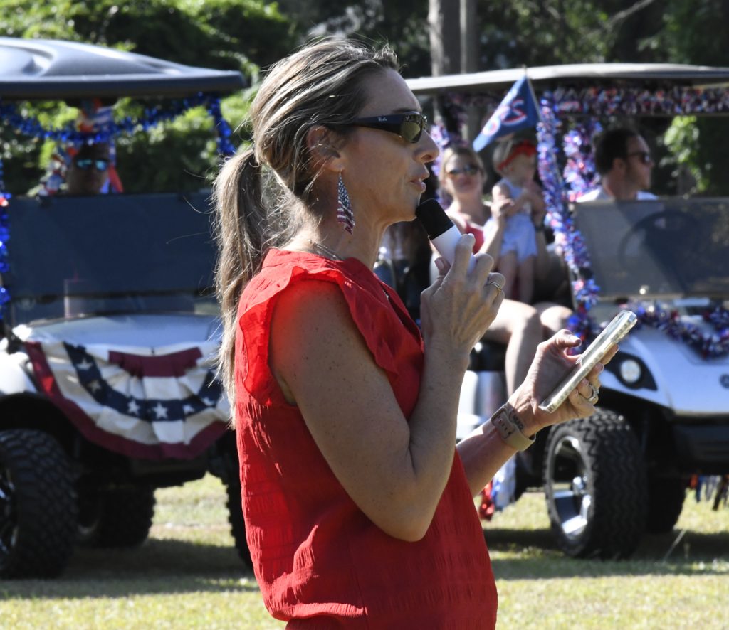 Woman in red top speaks into a microphone at an outdoor event, holding a phone, with decorated golf carts and people in the background.