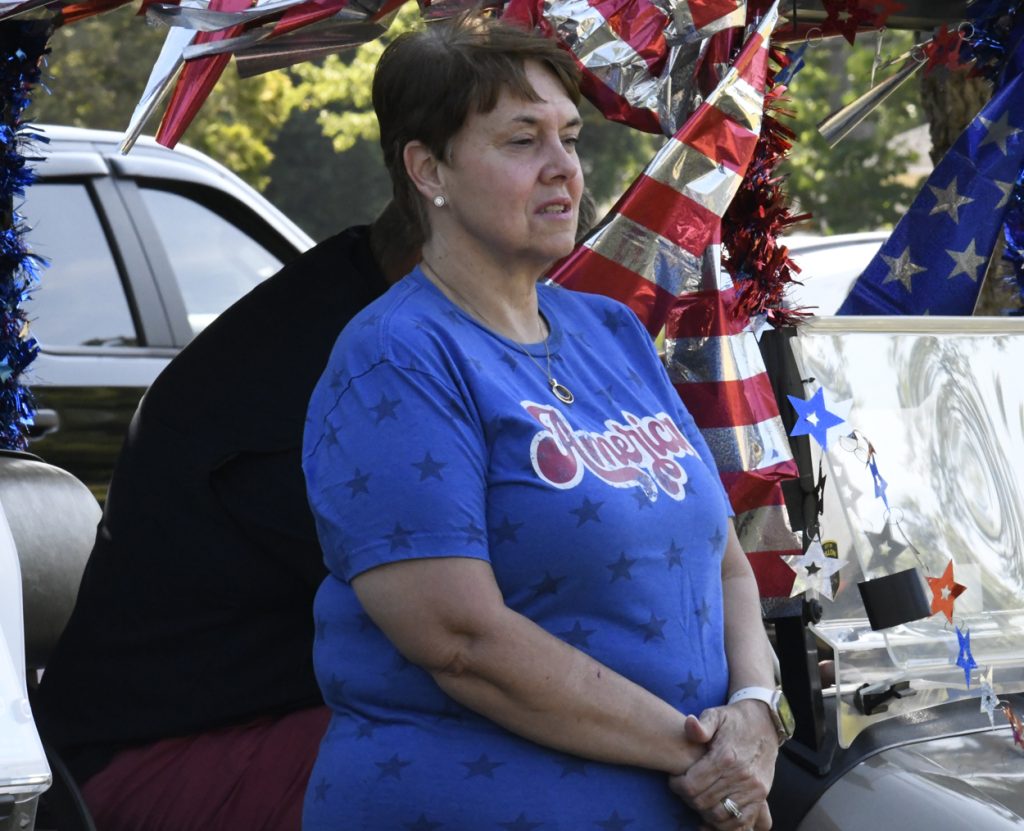 A woman in a blue "America" shirt stands beside a decorated vehicle with patriotic red, white, and blue decorations.