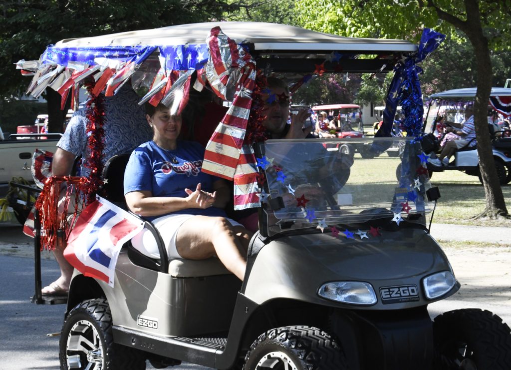 People riding a golf cart decorated with red, white, and blue patriotic streamers and flags for a parade or celebration, outdoors in a park setting.