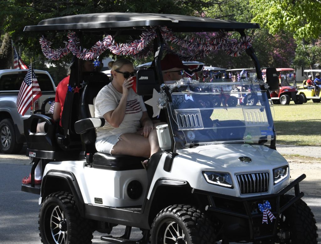 A decorated golf cart with American flags and tinsel, people riding inside, one woman in a patriotic shirt drinking from a red cup, outdoors during a parade.