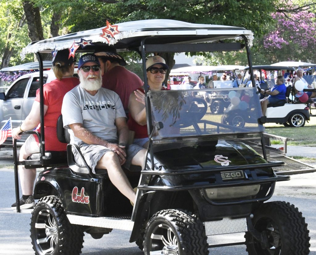 People riding a decorated golf cart during a parade, wearing patriotic hats and clothing, with American flags and festive decorations visible in the background.