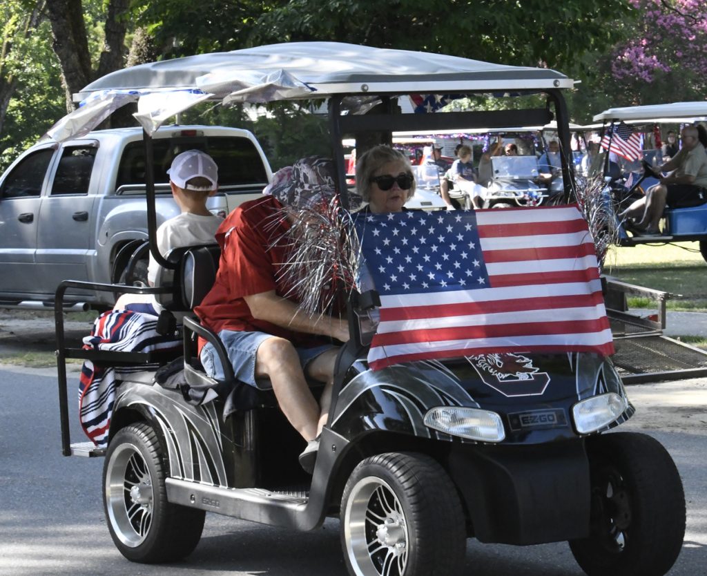 A golf cart decorated with an American flag and tinsel, driven in a parade with people inside and other decorated carts in the background.