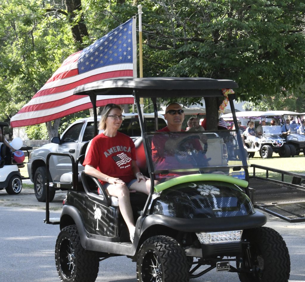 Two people ride a decorated golf cart with a large American flag during a parade, surrounded by trees, other carts, and people on a sunny day.