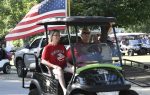 Two people ride a decorated golf cart with a large American flag during a parade, surrounded by trees, other carts, and people on a sunny day.