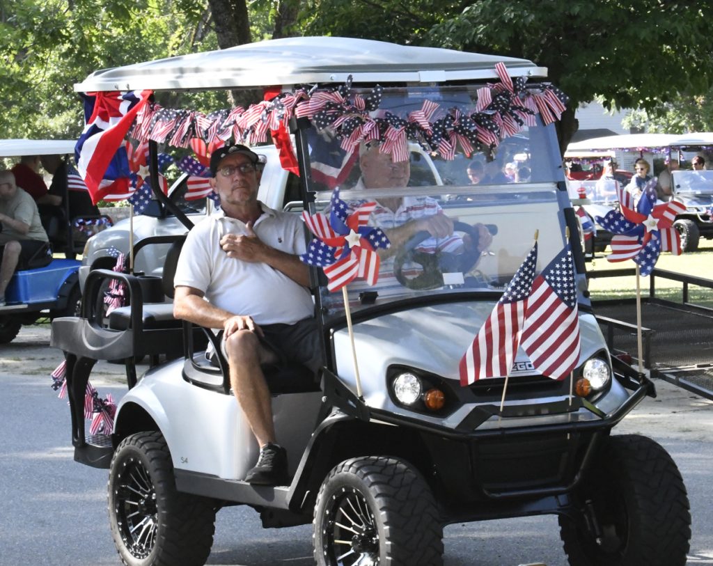 A golf cart decorated with American flags and pinwheels participates in a parade, with two men riding and spectators in the background.