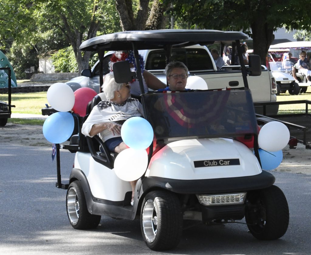 Two women riding in a decorated golf cart with red, white, and blue balloons on a sunny day in a park.