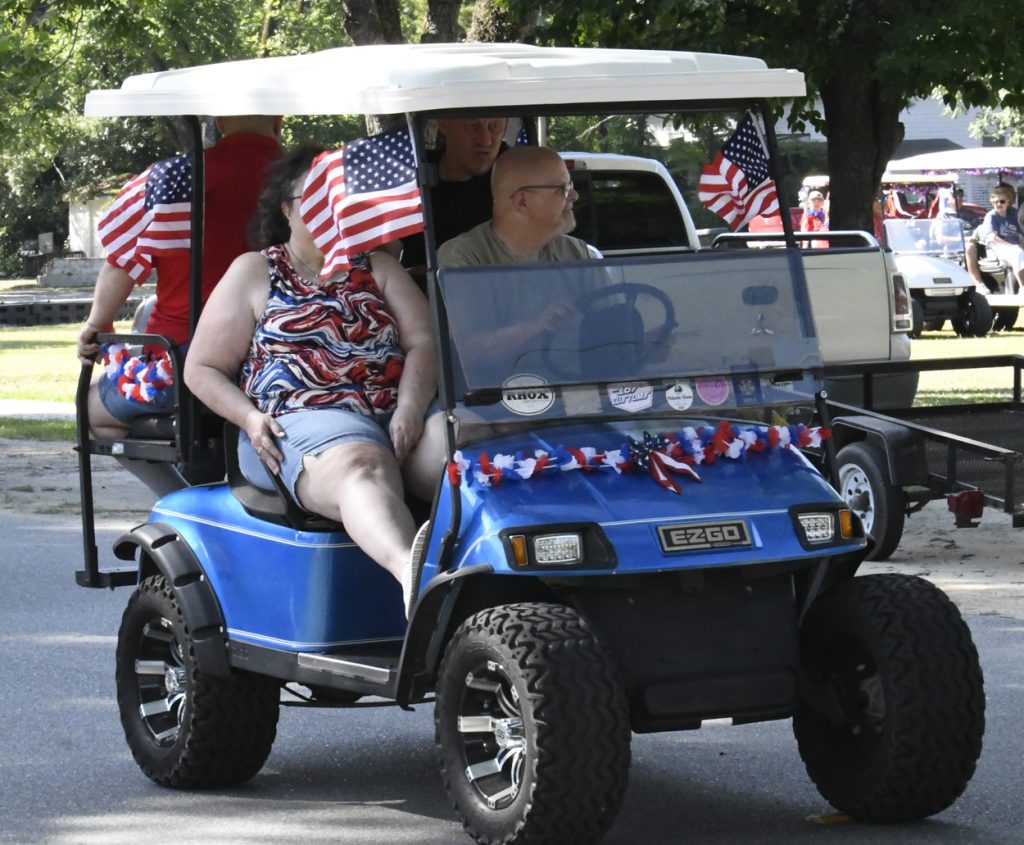 People riding a decorated blue golf cart with American flags during a parade, wearing casual summer clothing.