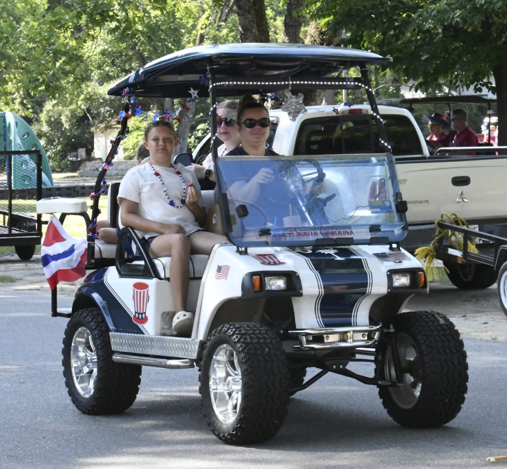 Three people riding a decorated golf cart with patriotic flags and garlands on a sunny day, with trees and parked vehicles in the background.