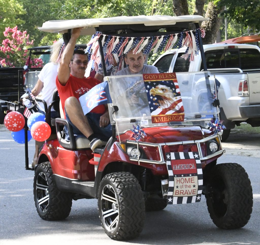 People riding a decorated golf cart with patriotic flags, balloons, and signs celebrating America, including "God Bless America" and "Home of the Brave," during a parade.