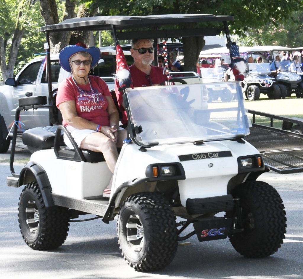 Two people wearing red shirts ride a decorated golf cart in a parade, with American flags and festive accessories, on a sunny day outdoors.