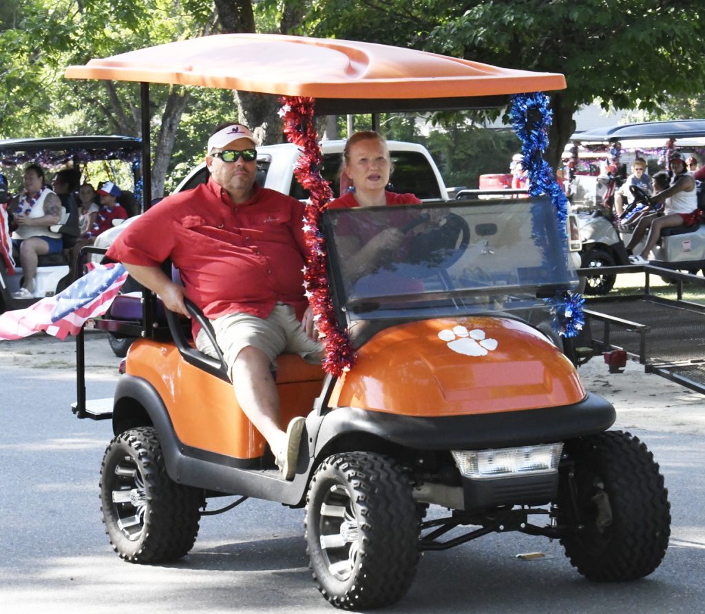 Two people ride an orange golf cart decorated with red and blue tinsel during a parade, surrounded by other decorated carts and spectators in the background.