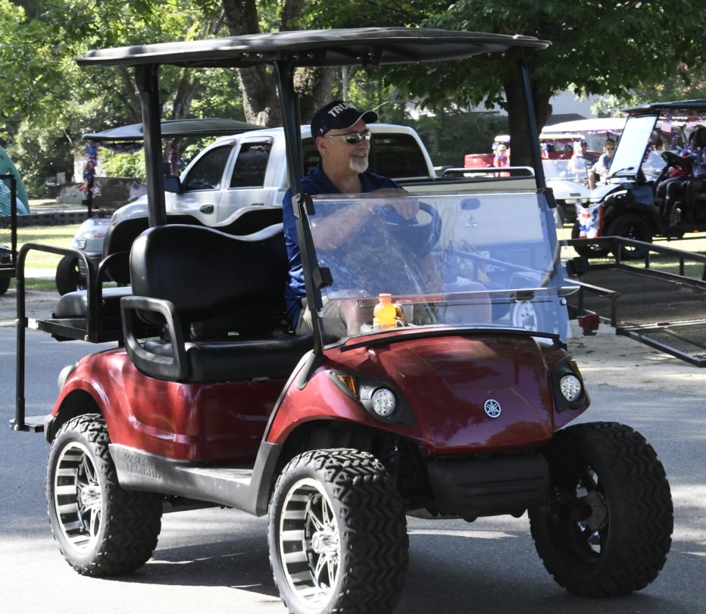 Man wearing sunglasses and a hat drives a red Yamaha golf cart outdoors on a sunny day, with other vehicles and trees in the background.
