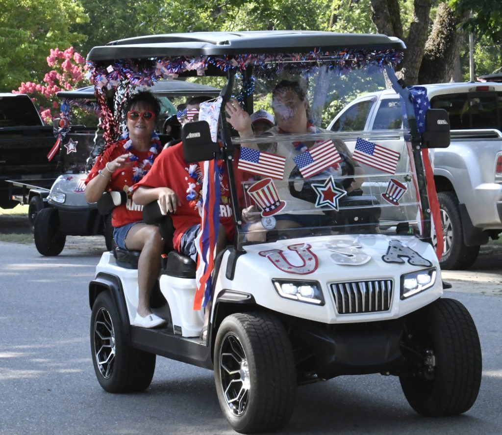 People riding a decorated golf cart with American flags and patriotic colors, celebrating outdoors on a sunny day.