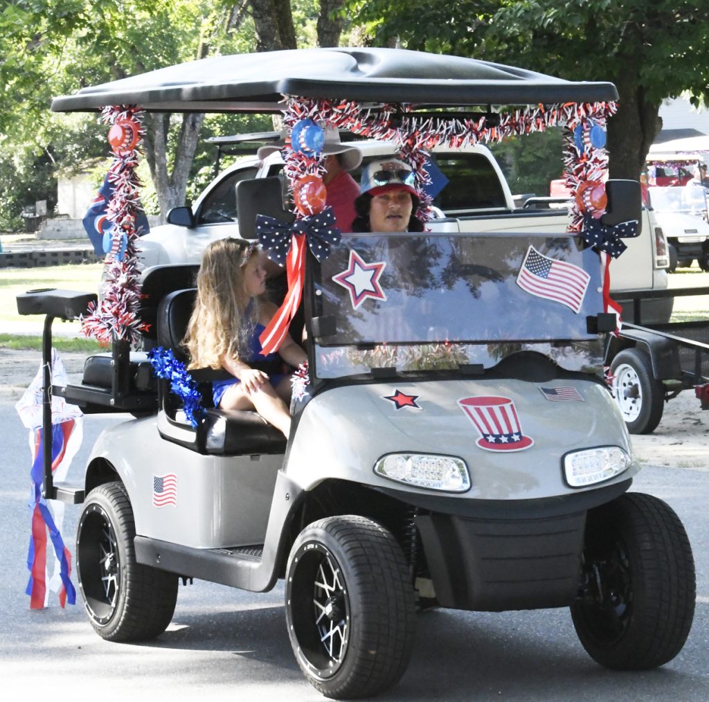 A golf cart decorated with patriotic red, white, and blue decorations, driven by a woman and carrying a child, during a parade or celebration.