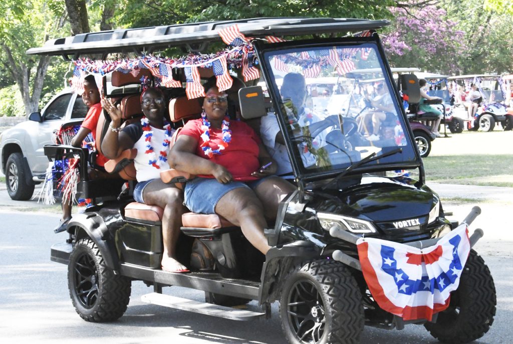 People riding a decorated golf cart with American flags and patriotic colors, waving and smiling during a festive parade on a sunny day.