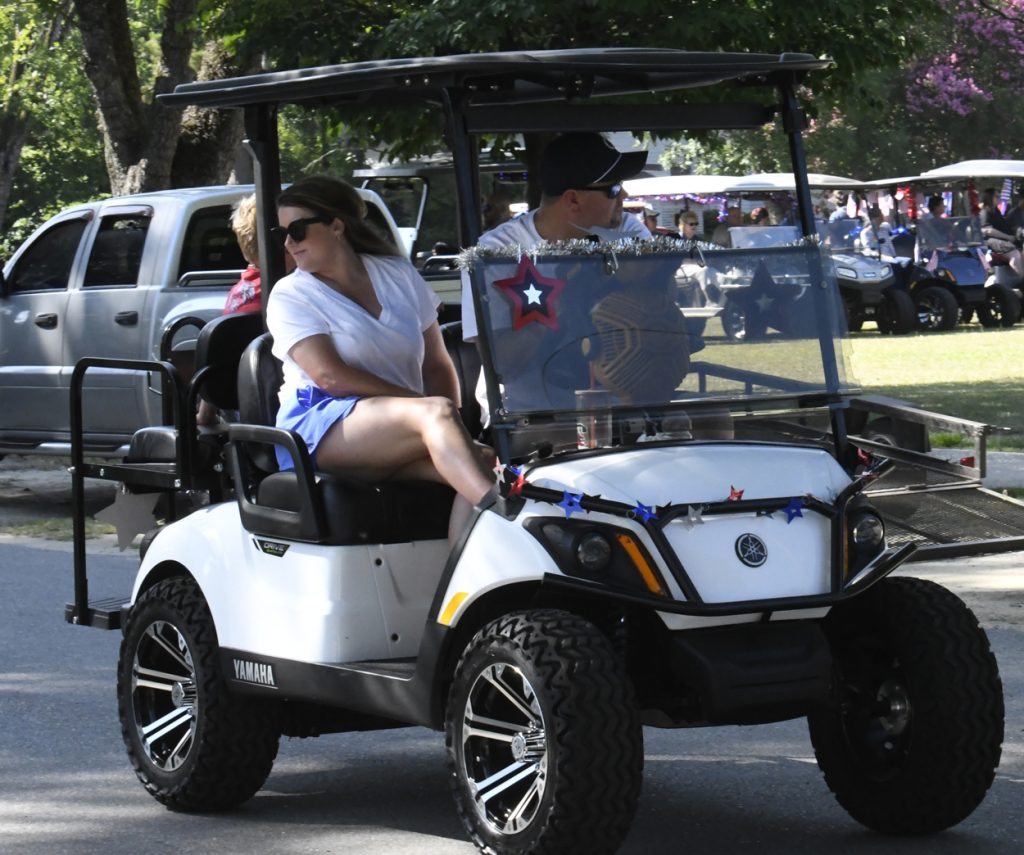 People riding a decorated golf cart on a sunny day, with red, white, and blue star decorations, parked vehicles, and other golf carts visible in the background.