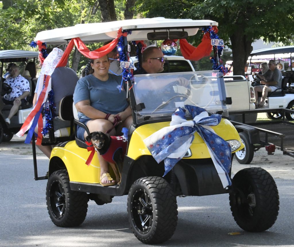 A yellow golf cart decorated with red, white, and blue ribbons and bows, carrying smiling people and a dog during a parade.