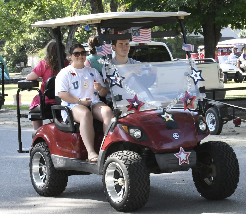 A decorated red golf cart with people riding, adorned with American flags and stars, participates in a parade on a sunny day.