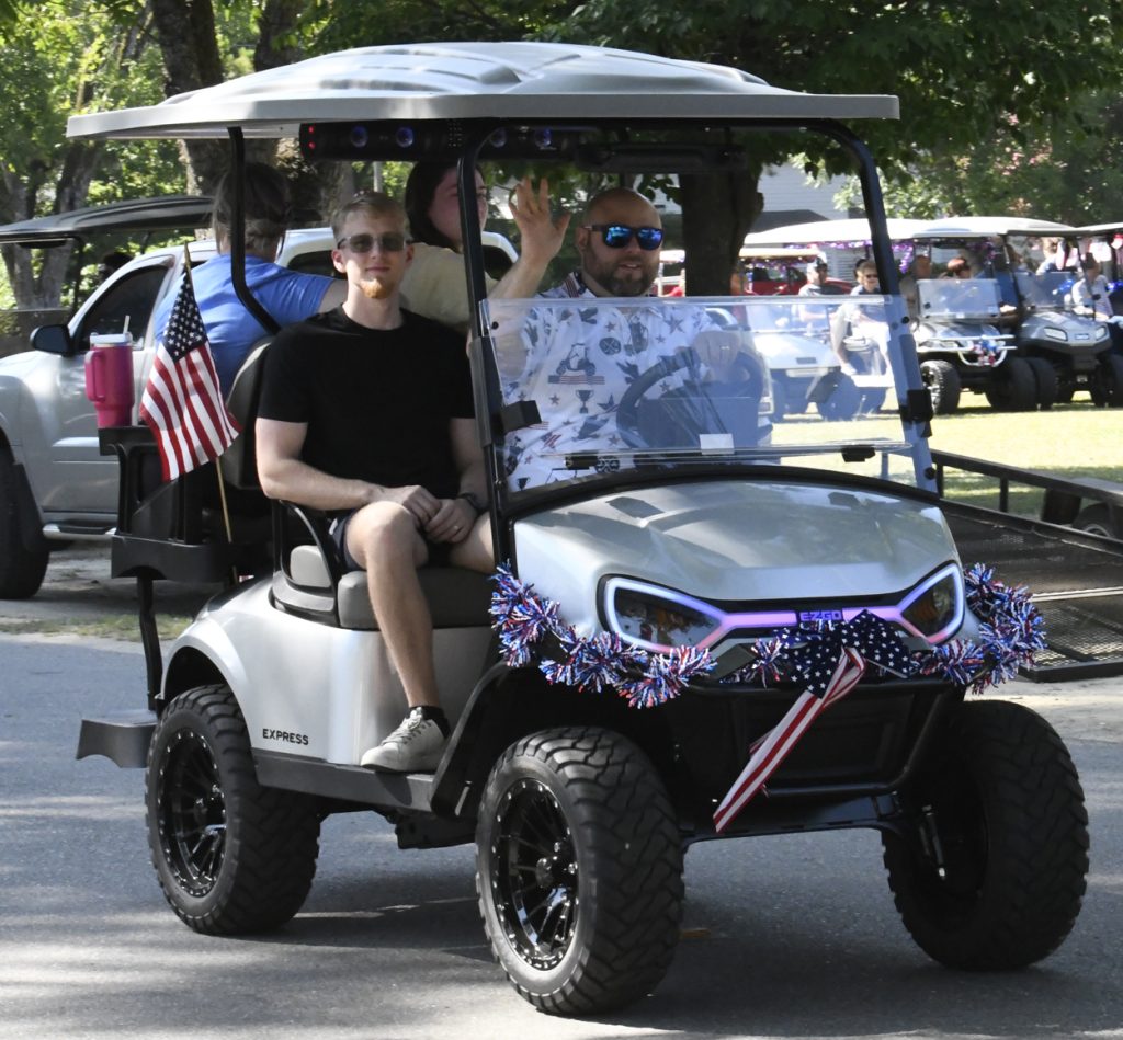 Two men ride a decorated golf cart with American flags and patriotic garlands during a parade, with trees and other golf carts visible in the background.