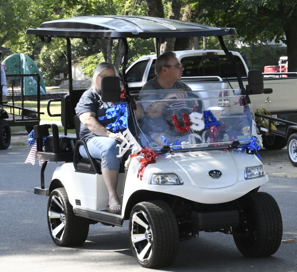 A decorated golf cart with "USA" on the windshield, driven by a man with a woman passenger, participating in a patriotic parade.