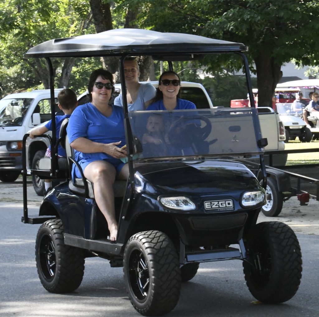 Four people ride a black EZGO golf cart on a sunny day, with trees and vehicles visible in the background.