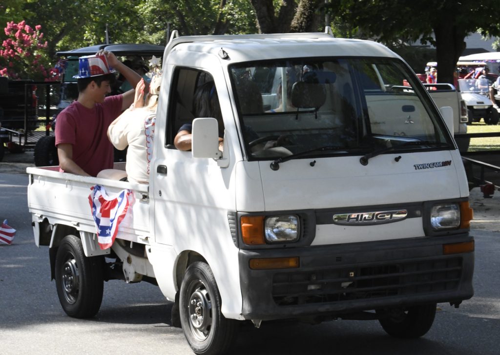 A small white pickup truck decorated with American flags and bunting, carrying people in festive hats during a daytime parade.