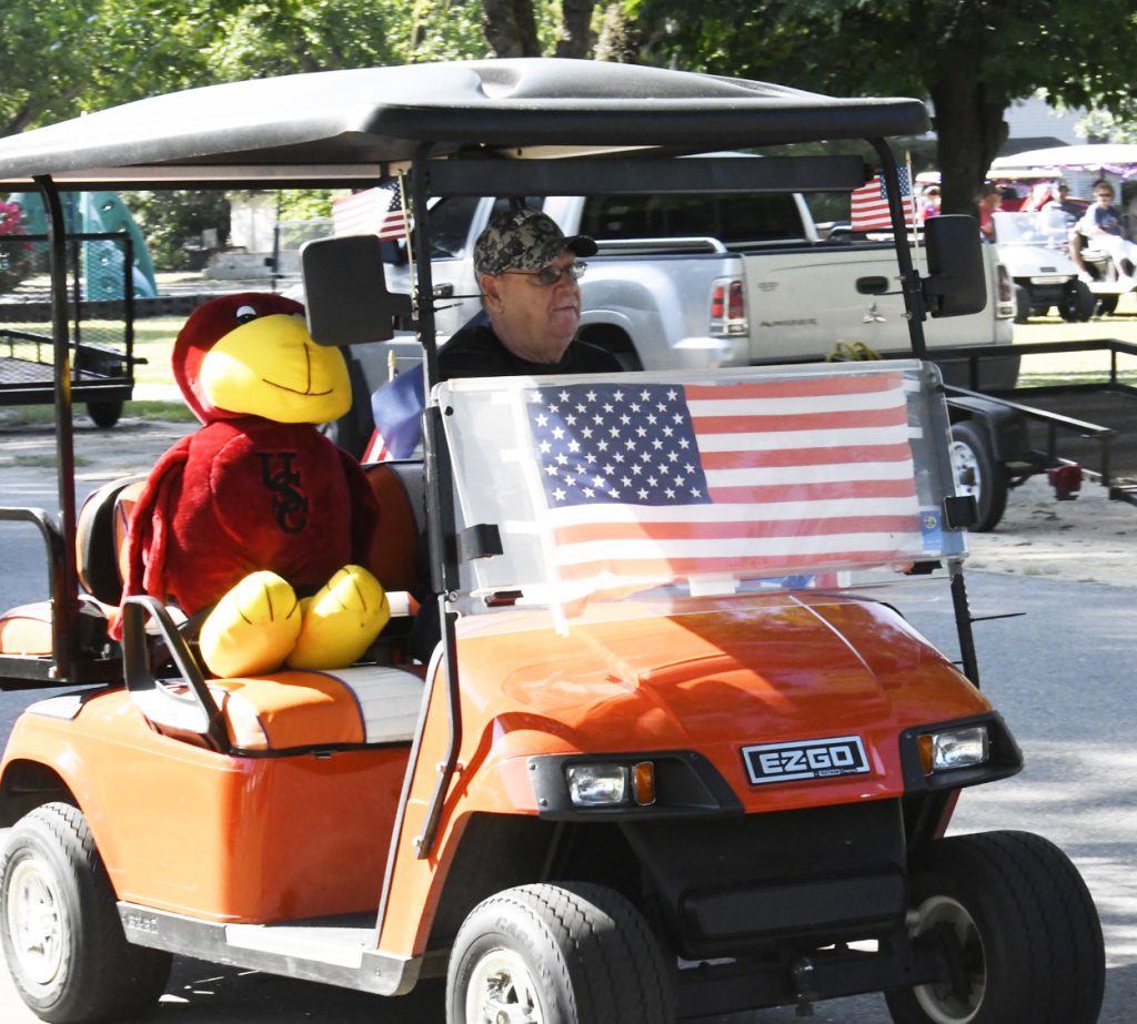 A man drives an orange golf cart decorated with an American flag, next to a large plush bird mascot, during a sunny outdoor event.