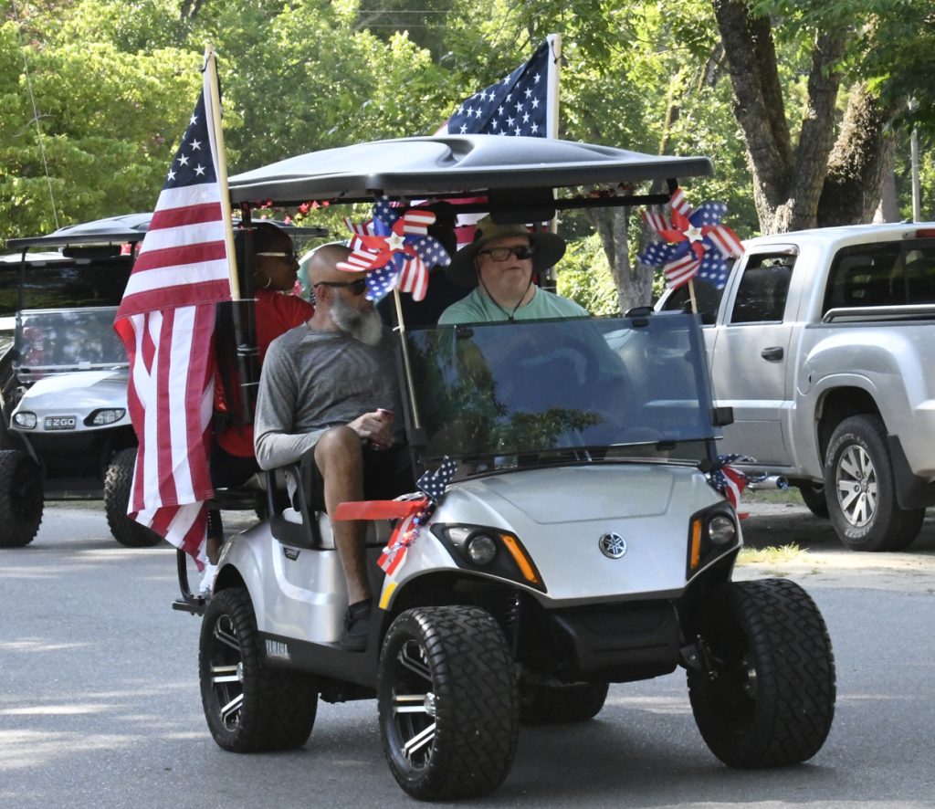 People ride a decorated golf cart with American flags and pinwheels in a parade, celebrating a patriotic event on a sunny day.