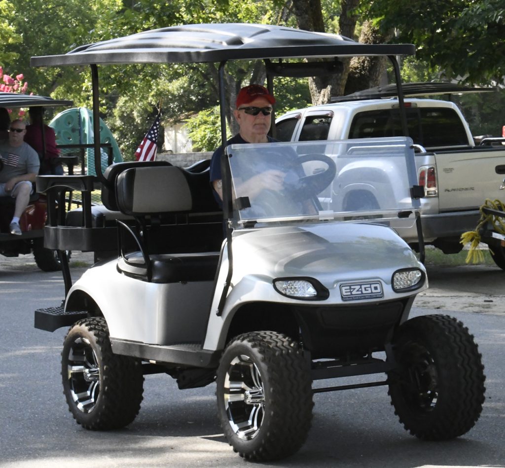 Man wearing a red cap and sunglasses drives a silver EZGO golf cart with an American flag attached, outdoors on a sunny day.
