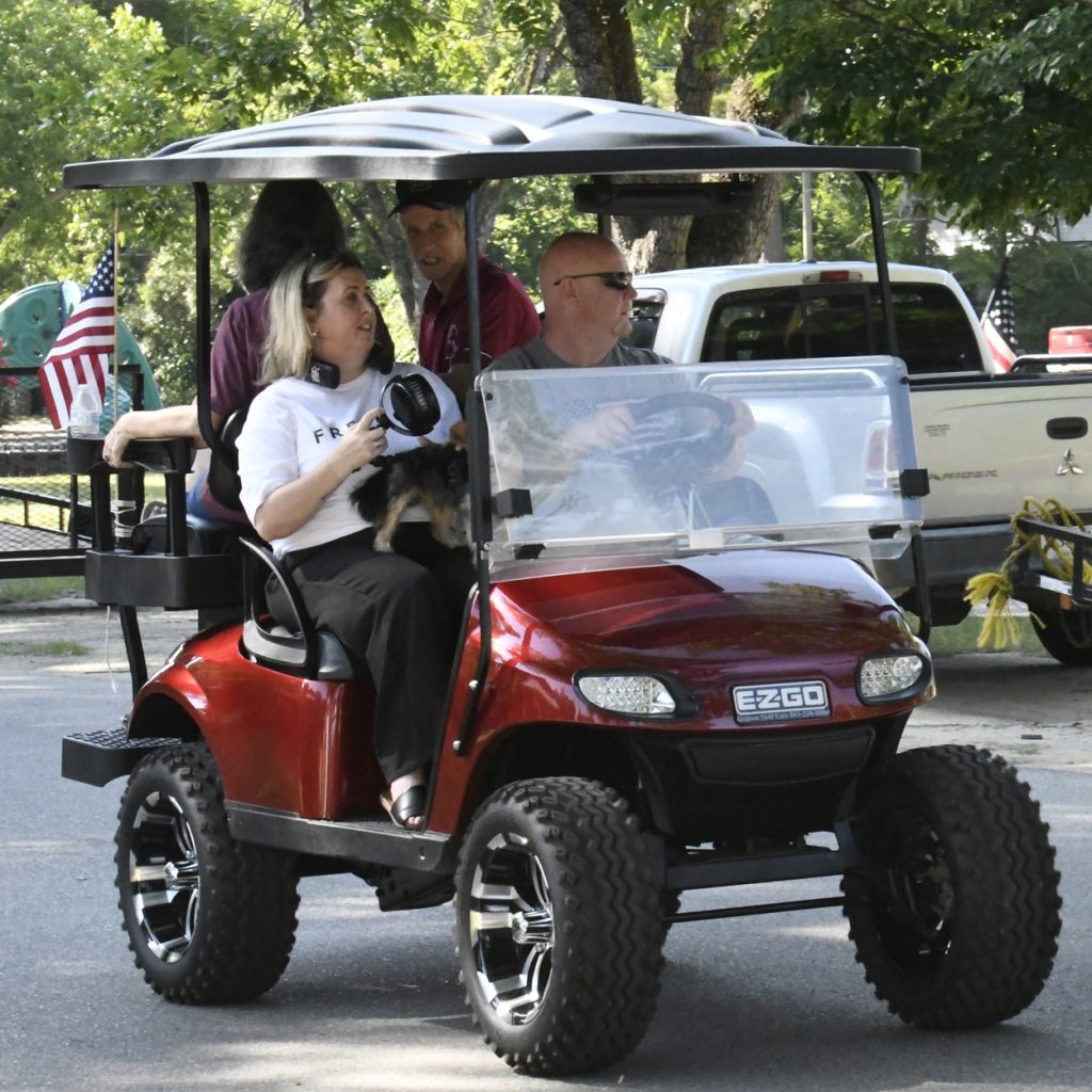 Four people and a small dog ride in a red EZGO golf cart, outdoors on a sunny day, with American flags visible in the background.